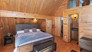 A cozy wooden cabin room with a bed, a bench, a small table with a plant, and a bathroom with a sink and toilet.