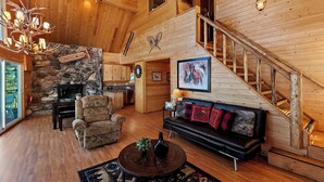 A spacious living area with a stone fireplace, wooden ceiling, and a staircase.