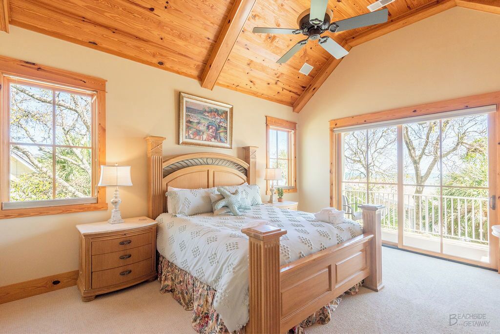 A bedroom with a wooden ceiling, a large bed with a headboard, two bedside tables, and a window with a view of trees.