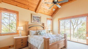 A bedroom with a wooden ceiling, a large bed with a headboard, two bedside tables, and a window with a view of trees. - 43 Dune Lane - 2nd Row Dune Lane Luxury, 100ft from the Sand (Hilton Head Island)