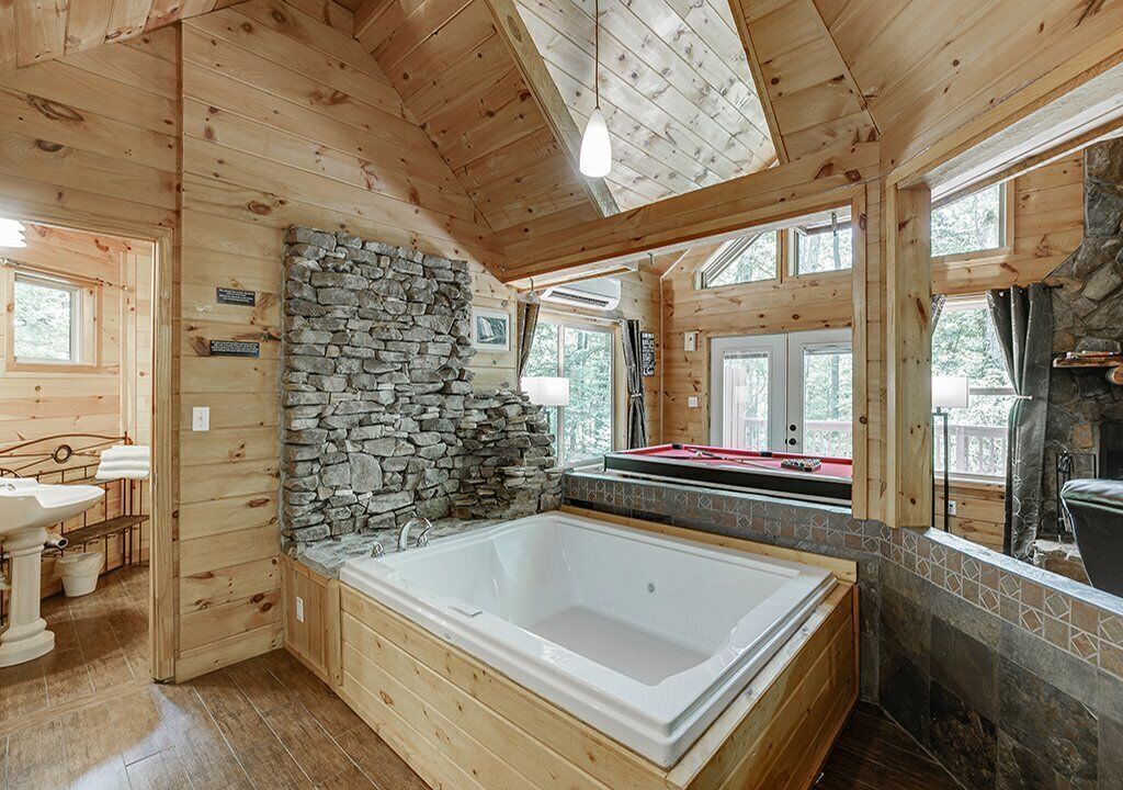 A log cabin bathroom with a stone wall, a large bathtub, wooden floors, and a stone fireplace.