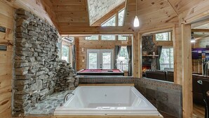 Interior de una cabaña de madera con pared de piedra, una bañera grande, mesa de billar y vistas al exterior.