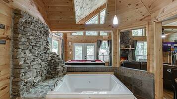 Interior de una cabaña de madera con pared de piedra, una bañera grande, mesa de billar y vistas al exterior.