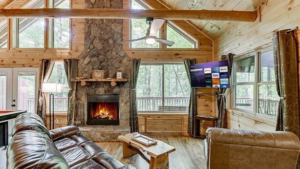 Un interior de cabaña acogedor con chimenea de piedra, sofás de cuero, una mesa de centro de madera y un televisor de pantalla plana montado en la pared.
