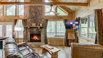 Un interior de cabaña acogedor con chimenea de piedra, sofás de cuero, una mesa de centro de madera y un televisor de pantalla plana montado en la pared.