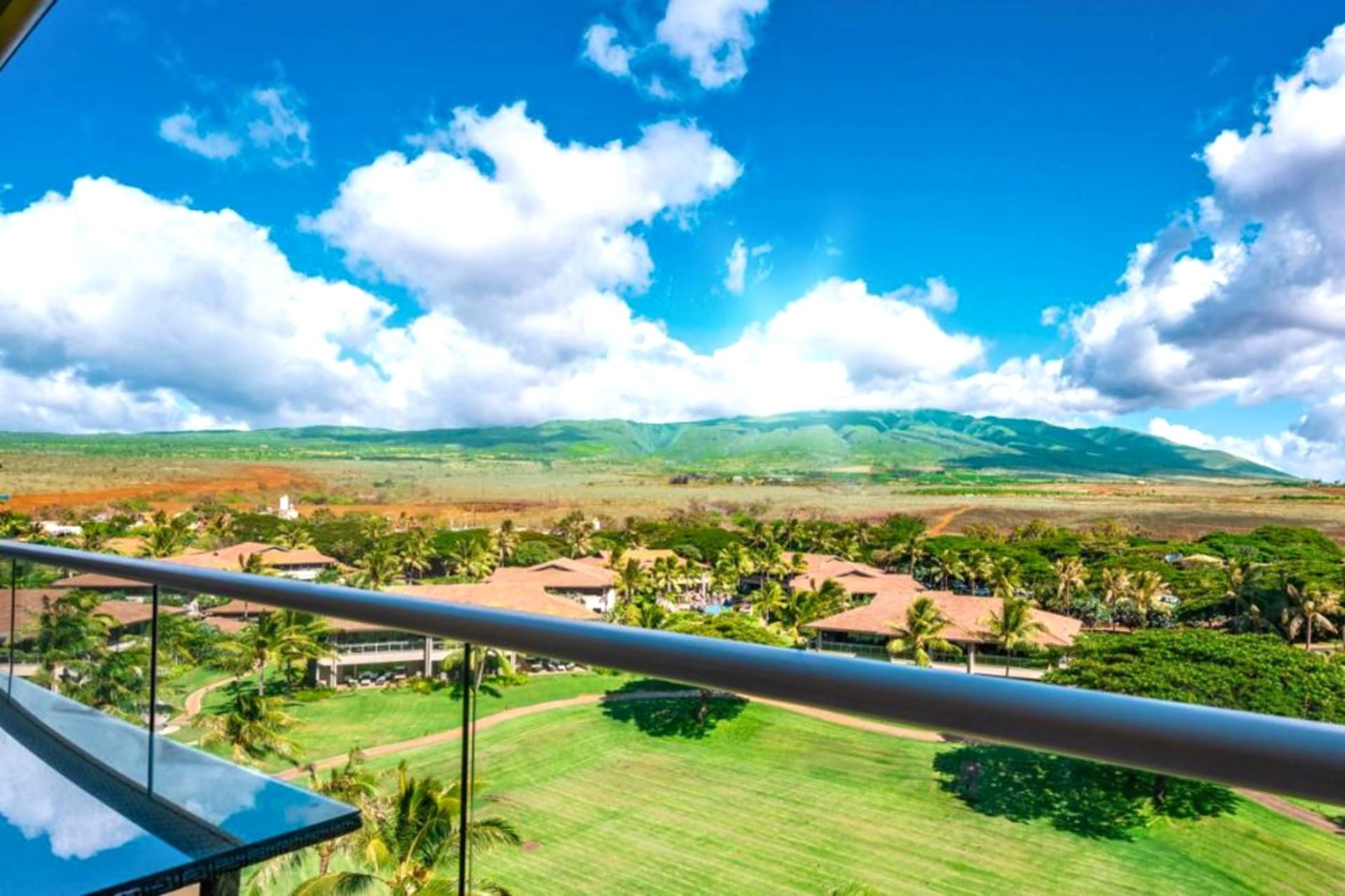 A view from a balcony overlooking a landscape with houses, palm trees, and distant mountains under a clear sky.