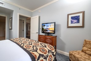A hotel room with a bed, a television on a wooden cabinet, a framed picture on the wall, and a door leading to another room.