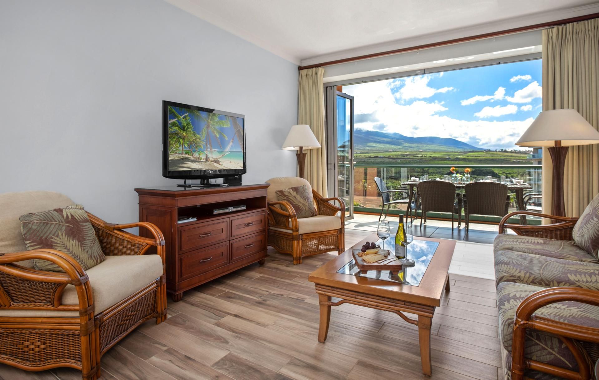 A living room with wicker furniture, a flat-screen TV, a glass-top coffee table, and a balcony with a scenic view.