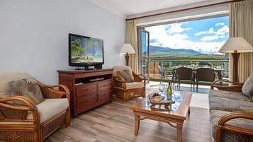 A living room with wicker furniture, a flat-screen TV, a glass-top coffee table, and a balcony with a scenic view.