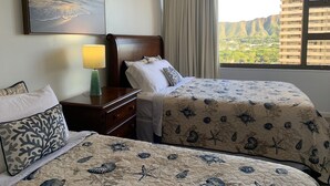 A hotel room with two beds, a nightstand, a lamp, a painting, and a window with a view of a cityscape. - Waikiki Banyan 1909 Amazing Views and Just  Steps to the Beach (Honolulu)