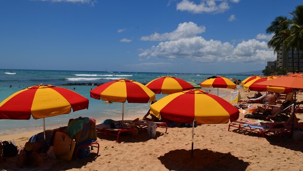 A beach with multiple red and yellow striped umbrellas, beach chairs, and a clear view of the ocean. - Waikiki Banyan 1909 Amazing Views and Just  Steps to the Beach (Honolulu)