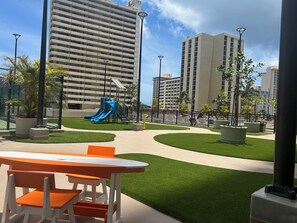 Un parque infantil con toboganes y un área cercada, rodeado de edificios y follaje.
