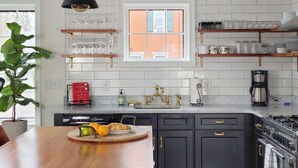 Una cocina moderna con azulejos blancos tipo metro, armarios negros y una encimera de madera. Se observan estantes con cristalería y un letrero que dice "BON APPETIT".