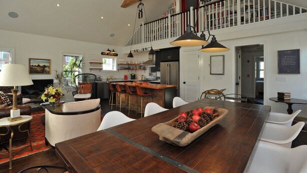 Amplia sala de estar con una gran mesa de comedor de madera, sillas blancas y una cocina al fondo.
