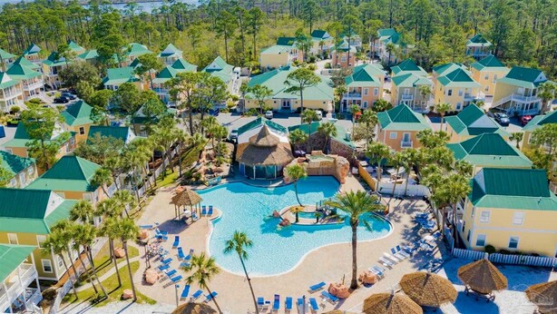Resort com vários edifícios, piscina central e palmeiras ao redor.