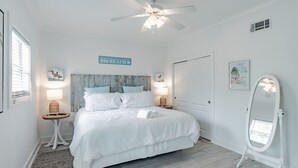 Quarto com cama, ventilador de teto, mesa de cabeceira com luminária, espelho fixado na parede e placa com tema praiano.