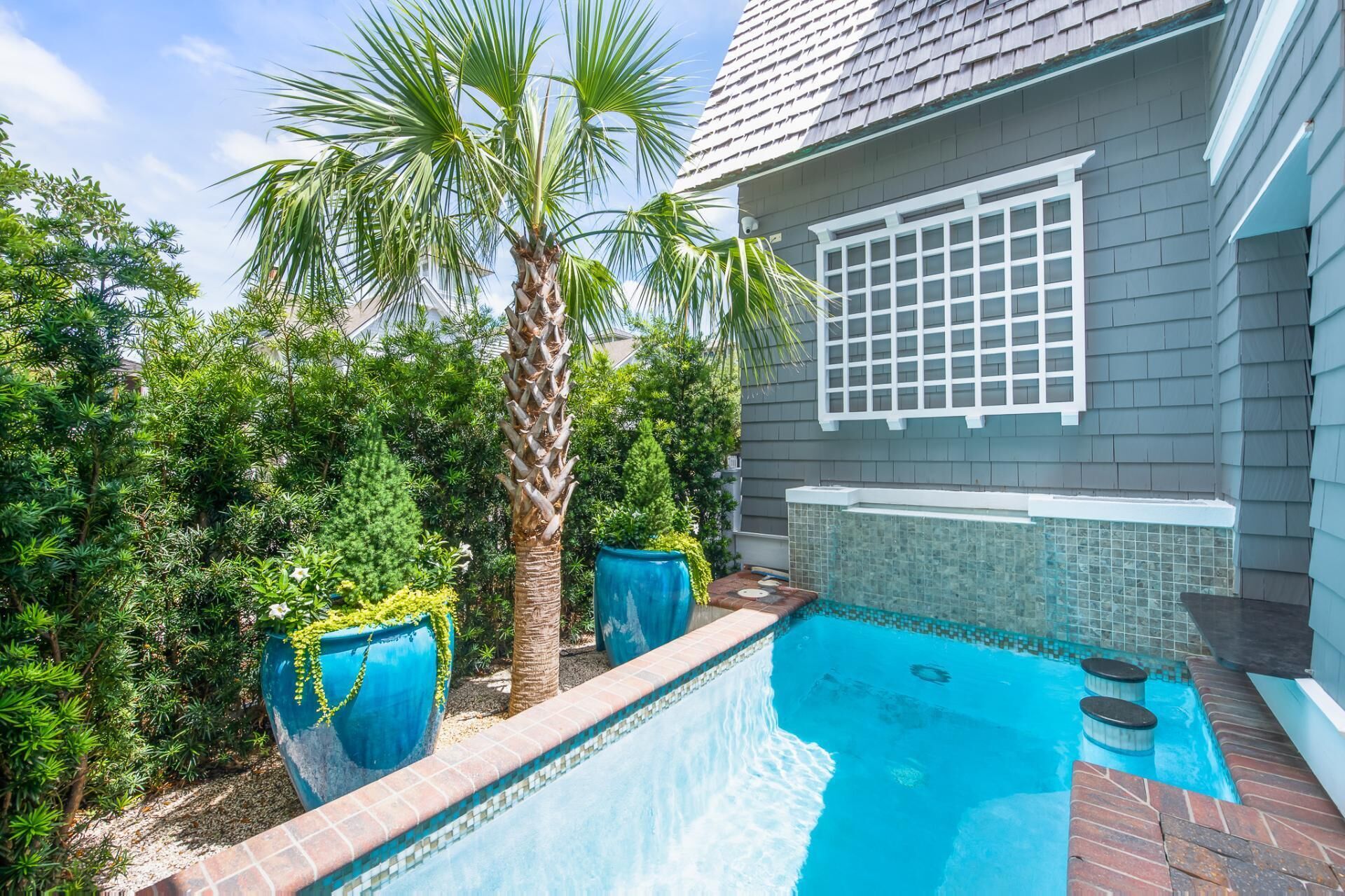 A swimming pool with a tiled edge, surrounded by lush greenery and a house with a grey exterior and a white window grille.