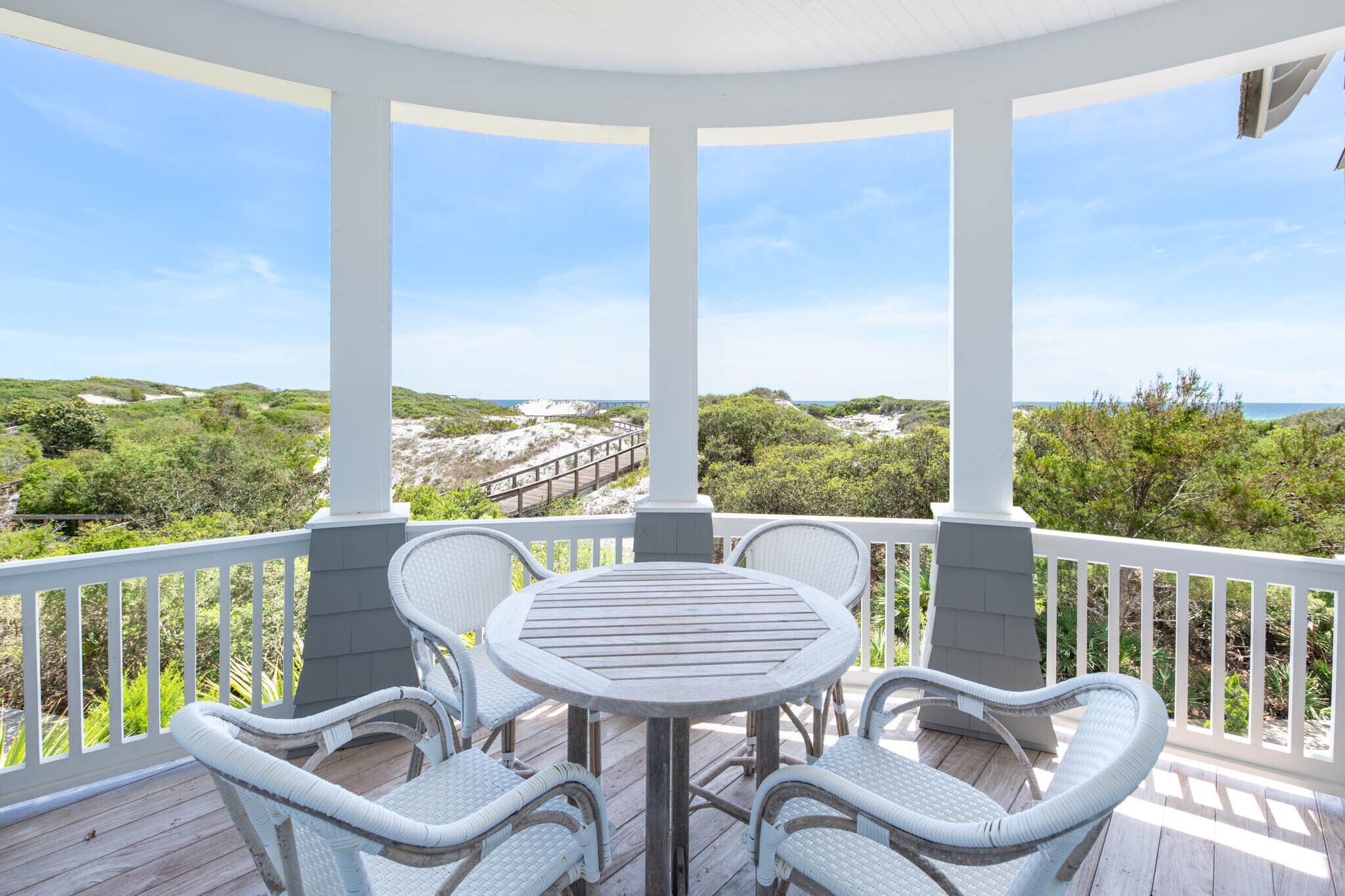 Een overdekt terras met een ronde tafel en rieten stoelen, met uitzicht op een kustlandschap met begroeiing en een zandstrand.