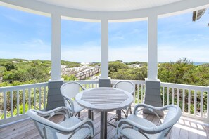 A covered outdoor area with a round table and wicker chairs, overlooking a coastal landscape with greenery and a sandy beach. - Beachfront with Pool and Golf Cart (Watersound)