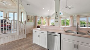 A spacious kitchen with white cabinets, a stainless steel dishwasher, and a large island with a sink. There are multiple windows with curtains, and a ceiling fan is visible. - Pelican Shores: Private pool, 4 bedrooms, Whalehead Beach (Corolla)