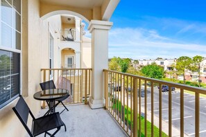 A balcony with a table and chairs, a view of a parking lot, and a building with multiple balconies.