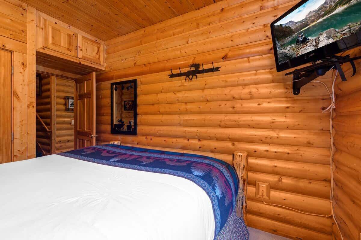 A log cabin room with a bed, a mounted television, and a wooden ceiling.