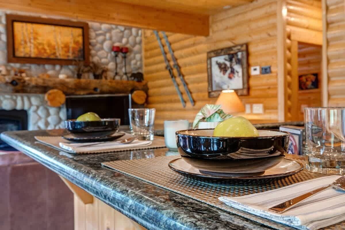 A wooden interior with a stone fireplace, a dining table with a bowl of fruit, and a lamp.