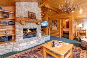 L’intérieur d’une cabane en rondins, avec une cheminée en pierre, des étagères en bois, un téléviseur et une table sur laquelle sont posées des bouteilles.