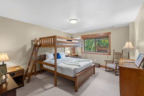A bunk bed room with a ladder, a desk, a chair, a lamp, and a window with a view of trees.