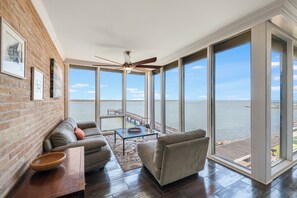 Una habitación amplia con un gran ventanal con vista al agua, un ventilador de techo, un sofá y una mesa de centro.
