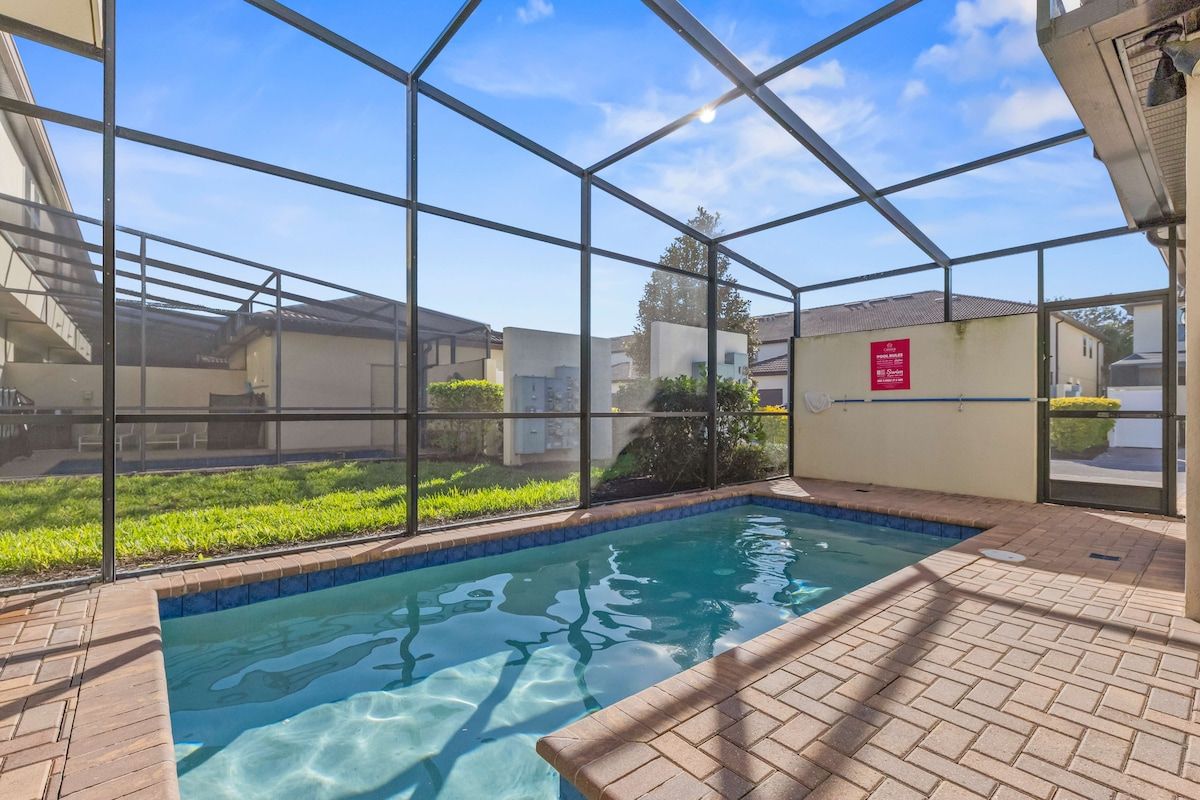 Piscina com proteção de vidro, cercada por um terraço de tijolos e vista para edifícios próximos.