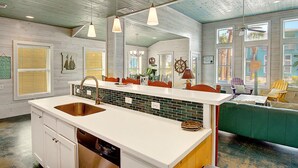 A kitchen with a large island, white countertops, and a green-tiled backsplash. There are pendant lights, a ceiling fan, and a view of the outside through large windows.