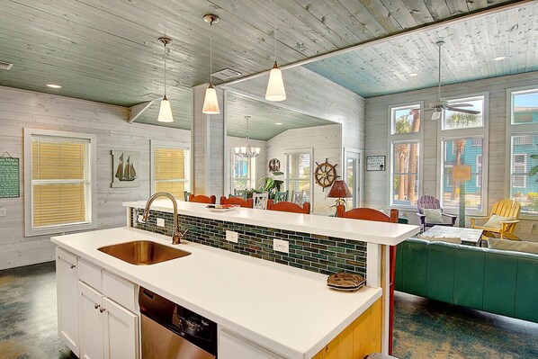 Una cocina con una isla grande, encimeras blancas y un salpicadero de azulejos verdes. Hay luces colgantes, un ventilador de techo y vistas al exterior a través de amplias ventanas.