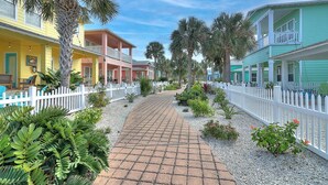 A pathway lined with white picket fences, leading to colorful buildings with balconies.