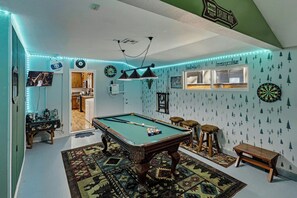 Une table de billard dans une pièce dont le plafond est vert, avec un jeu de fléchettes et une horloge murale.