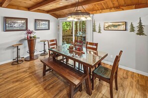 A dining area with a glass-top table, wooden chairs, a bench, and a hanging light fixture. - Outdoor Living by Snow Summit! Big Bear Mural! PET FRIENDLY! HOT TUB! Fire Pit! Large Fenced Yard! (Big Bear Lake)