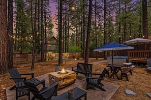An outdoor seating area with Adirondack chairs, a fire pit, and a wooden table. - Outdoor Living by Snow Summit! Big Bear Mural! PET FRIENDLY! HOT TUB! Fire Pit! Large Fenced Yard! (Big Bear Lake)