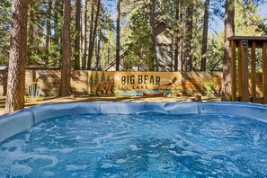 A hot tub surrounded by a wooden deck, with a sign reading 'Big Bear Lake' in the background. - Outdoor Living by Snow Summit! Big Bear Mural! PET FRIENDLY! HOT TUB! Fire Pit! Large Fenced Yard! (Big Bear Lake)