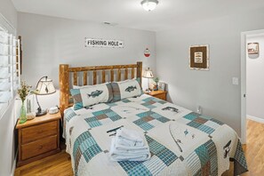 Une chambre à coucher avec une tête de lit en bois, une courtepointe ornée de motifs de poissons, une lampe de chevet, un tableau encadré et un panneau indiquant un endroit où l’on peut pêcher.