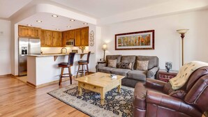 A modern kitchen with wooden cabinets, a central island, and bar stools. There is a living area with a sofa, a coffee table, and a patterned rug. - Beautiful 1 bedroom, 1 bath condo on Lake Dillon in Frisco. Walk to shuttle. (Frisco)