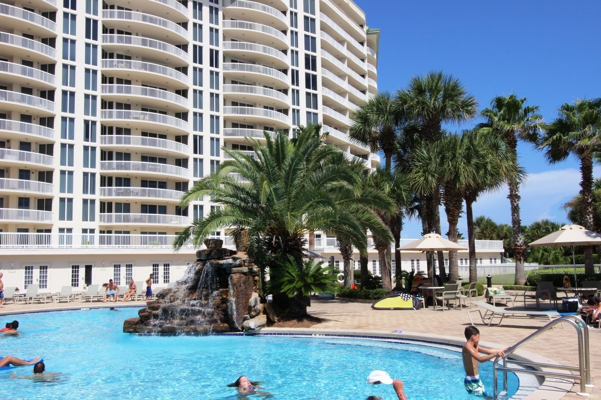 A hotel with a large pool featuring a waterfall, surrounded by palm trees and lounge chairs.