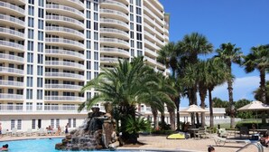 A hotel with a large pool featuring a waterfall, surrounded by palm trees and lounge chairs.