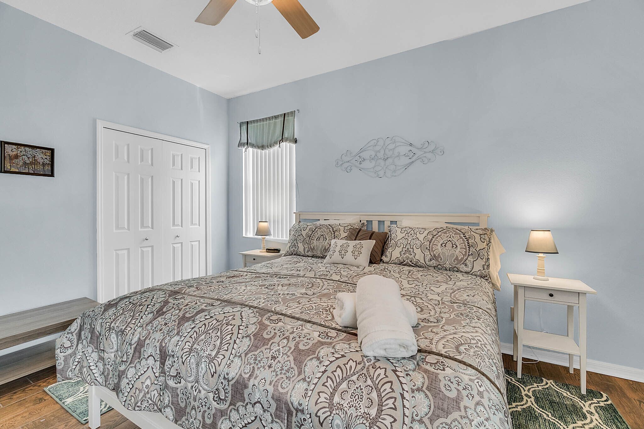 A bedroom with a bed, a nightstand, a ceiling fan, a window with curtains, and a wall decoration.