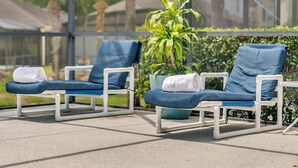 Dos sillones de descanso con cojines azules y estructura blanca, ubicados en un patio de concreto con una planta grande en maceta entre ellos.
