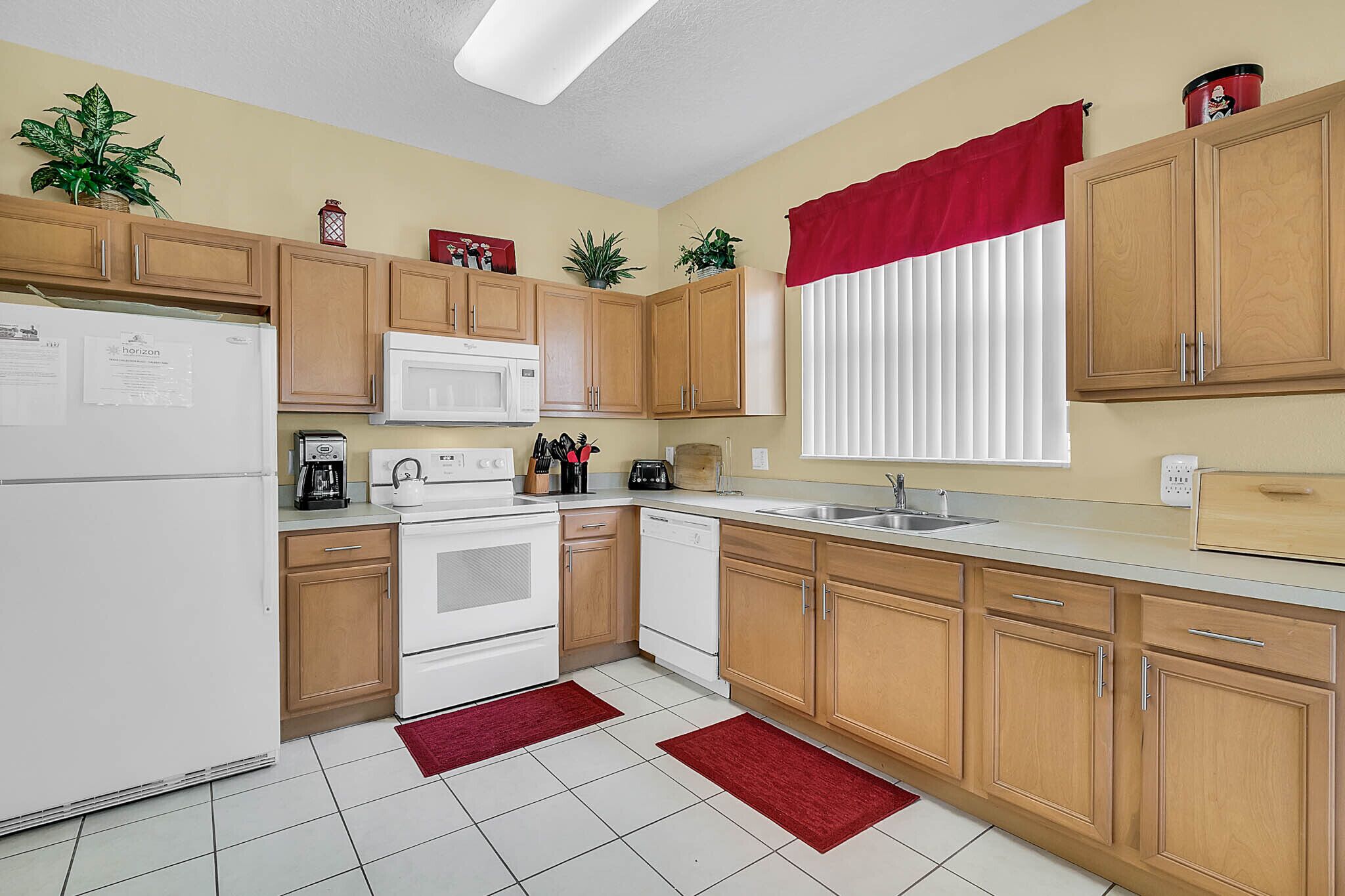 A compact kitchen with wooden cabinets, a white refrigerator, a microwave, a stove, and a dishwasher. There are two red rugs on the floor.