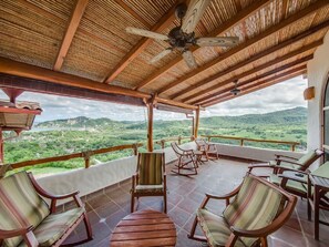 Relax on the balcony with a view of lush green surroundings, complete with a seating area and ceiling fan.