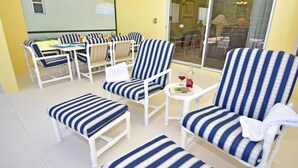 Patio soleggiato con arredi a righe bianche e blu, tra cui sedie, pouf e un tavolino rotondo con una bottiglia e un bicchiere.
