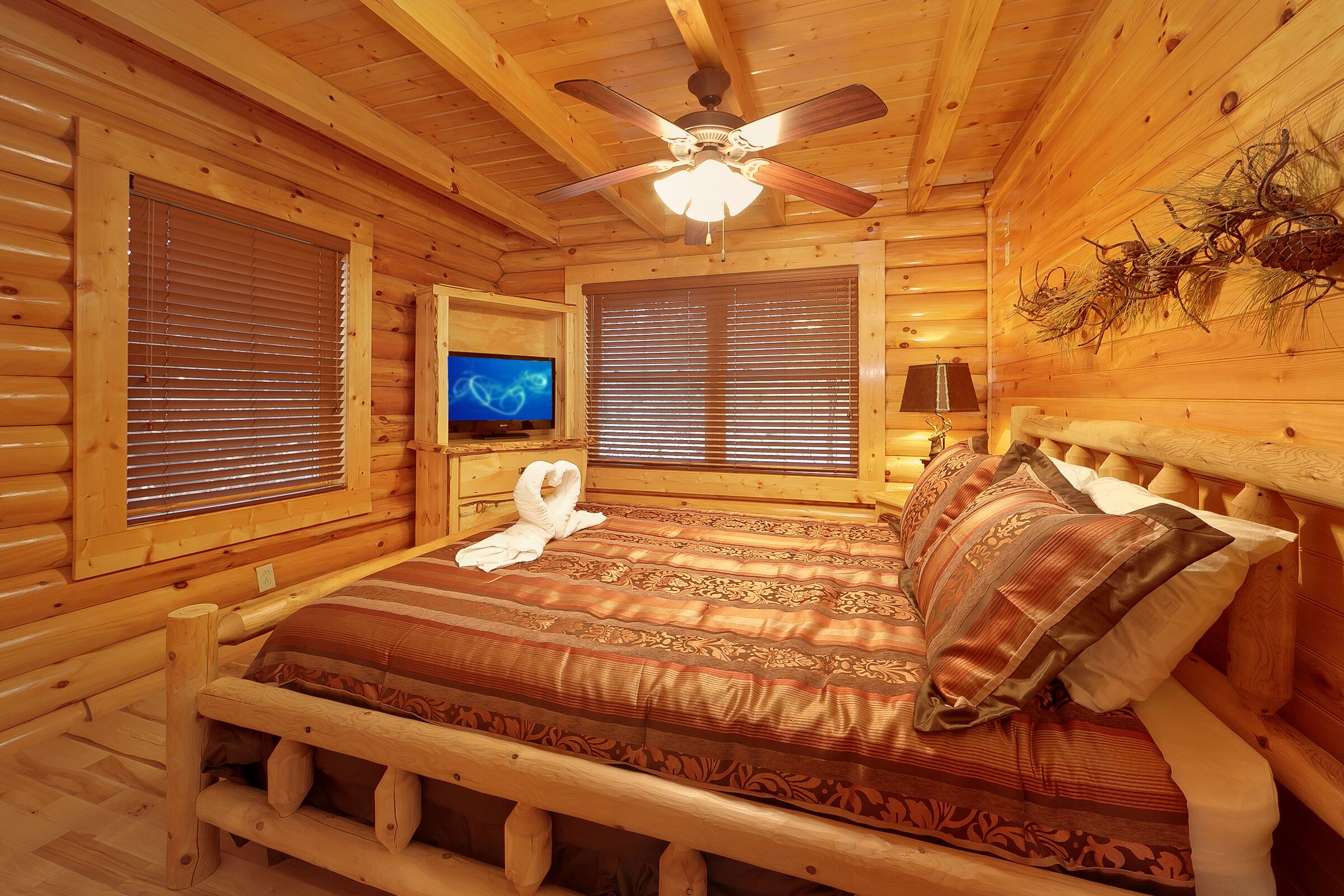 A log cabin bedroom with a wooden bed, two windows with blinds, a ceiling fan, a television on a stand, and a wall-mounted lamp.