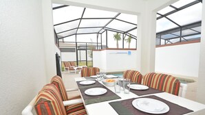 A covered outdoor dining area with white tables and striped cushioned chairs, a pool in the background, and a clear view of the sky. - Stunning Town Home with Splash Pool in Champions Gate Resort - Close to Theme Parks (Davenport)
