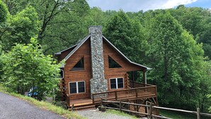 Eine Holzhütte mit einem Steinsschornstein, einer Holzdachterrasse und Blick auf den umliegenden Wald.